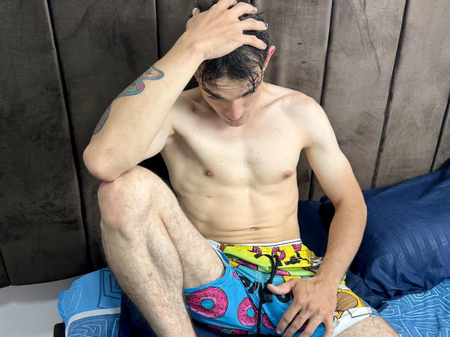 Keytwoo sem camisa, com um corpo esguio e musculoso, com abdominais visíveis, vestindo shorts coloridos com estampas de donuts, sentado em uma cama com um travesseiro azul, com a mão no cabelo, olhando para baixo..