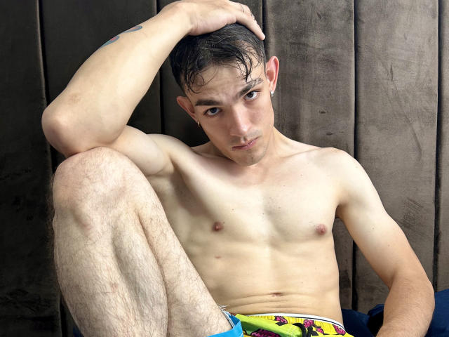 Keytwoo, sem camisa, com cabelo curto e escuro, pequenos mamilos e uma tatuagem no braço, está sentado com uma perna levantada, vestindo roupa íntima colorida, olhando para a câmera com um leve sorriso..