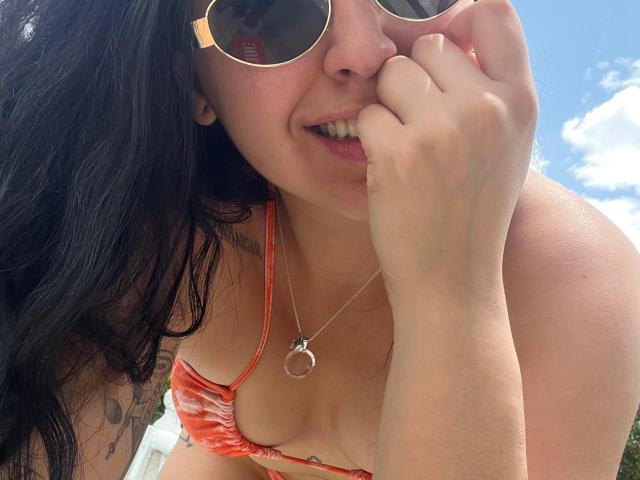 MalenaScordia with dark hair, wearing orange bikini top, showing her tits, holding her finger near her mouth, wearing sunglasses, necklace with ring pendant, smiling, under bright sunlight.