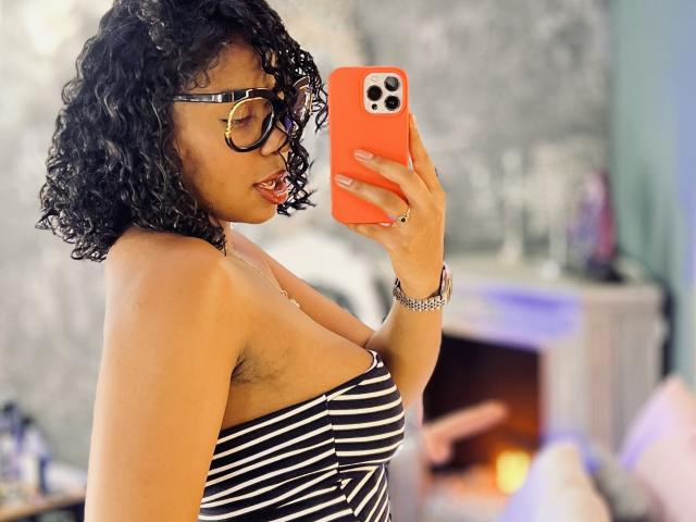 LisaMendez with curly black hair, glasses, and a striped black-and-white top, taking a selfie with an orange phone, showing her small tits and armpit hair.
