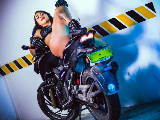 ZaraBeicker with black hair, red lipstick, and tattoos, sitting on a black motorcycle, legs raised, wearing a black leather outfit, gloves, and boots, in a blue-lit garage.