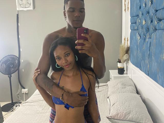 CataleyaAndEthan, une femme noire aux longs cheveux noirs, portant un haut floral bleu, embrassant un homme noir grand, en chemise noire, avec un pantalon déchiré, tenant un téléphone, devant un mur bleu avec un motif doré, avec un lit blanc..