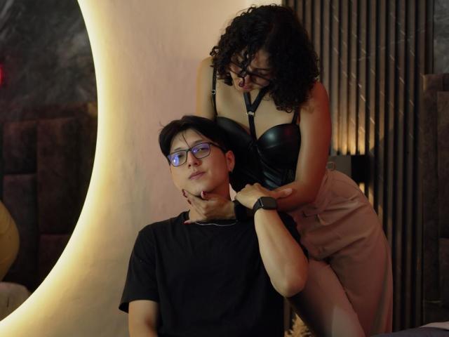 VanillaHotG with curly brown hair and black lingerie, her tits pressed against a wall, being kissed by a shirtless VanillaHotG with glasses and short black hair.