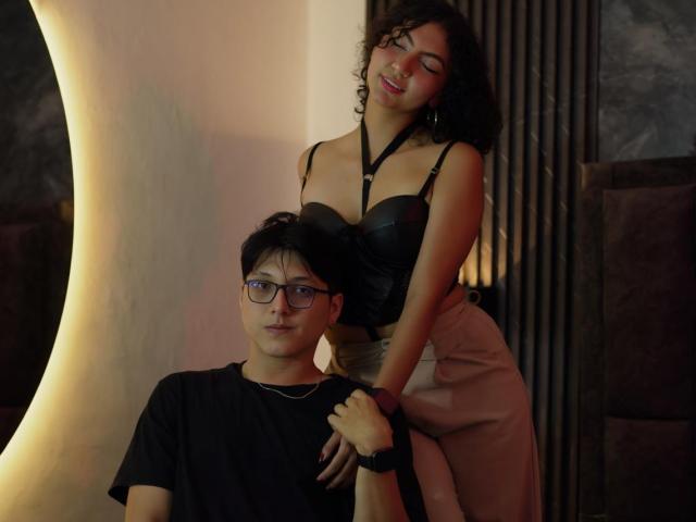 VanillaHotG with curly black hair and tan skin, wearing a black leather top, kisses a dark-haired man in a black shirt. His hands are on her ass.