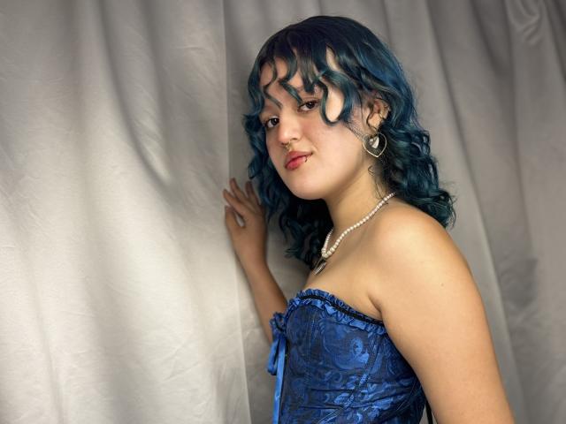 StephyStar, com cabelo encaracolado azul, vestindo um corpete de renda azul, um colar de pérolas e brincos de coração, está em pé em frente a uma cortina branca, tocando-a com a mão direita..