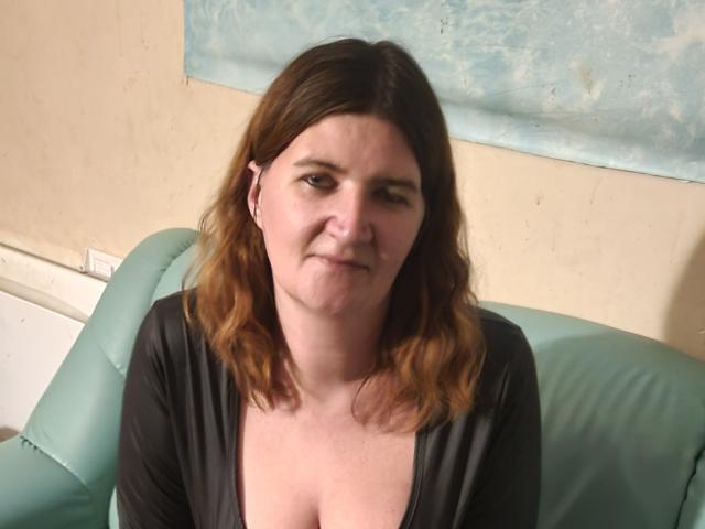 LeilaCrystalx, met halflang bruin haar, een lichte glimlach en kleine borsten, draagt een zwarte top en zit op een groene leren bank, waarbij ze recht in de camera kijkt..