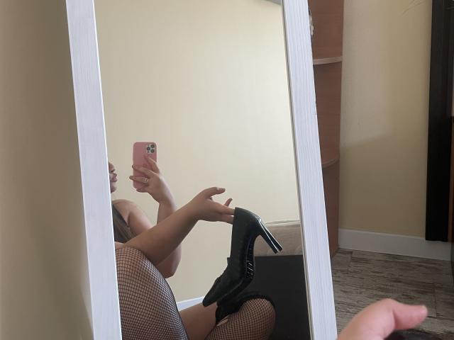 Madrinia's reflection in a mirror shows her taking a selfie with a pink phone, wearing fishnet stockings and black high heels, holding the heels in her right hand.