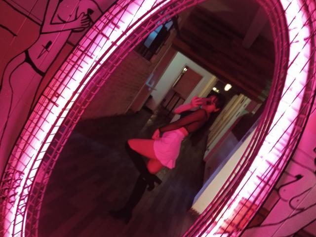 MiaPoe, vestita con un abito rosa, stivali neri e una maglietta rossa, è in posa con il sedere rivolto verso uno specchio con cornice rosa in una stanza poco illuminata. Il suo seno è piccolo..