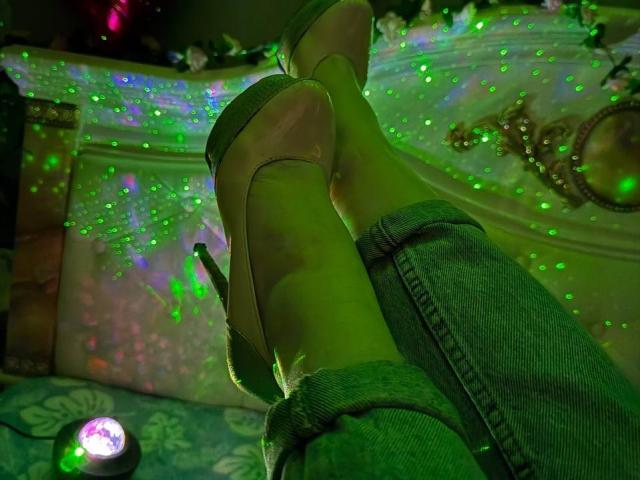 Siyana's groene, lichtgevende voeten in hoge hakken, gekruist bij de enkels, draagt blauwe jeans, met een kleine spotlight op het bed. Glinsterende groene lichten en een sierlijke ketting op de achtergrond..