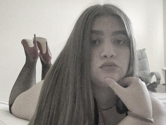 Madrinia with long brown hair, lying on a bed, legs up, wearing black fishnet stockings and red high heels, hand under chin, looking serious, in grayscale.