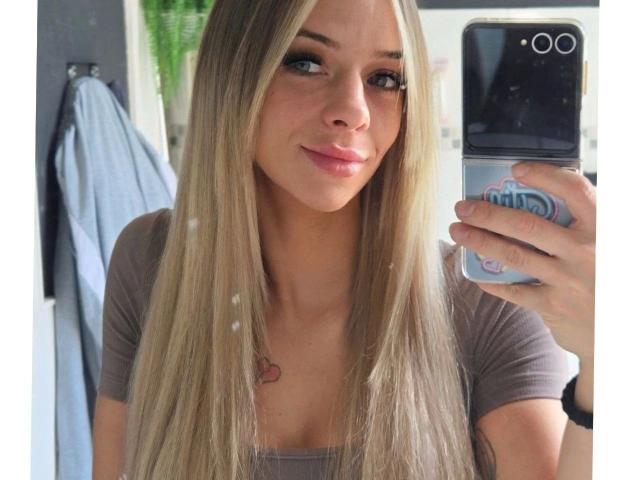 LexaXoXo-hot with long blonde hair, brown eyes, and a small heart tattoo on her chest, taking a selfie with a blue pop socket on her phone.