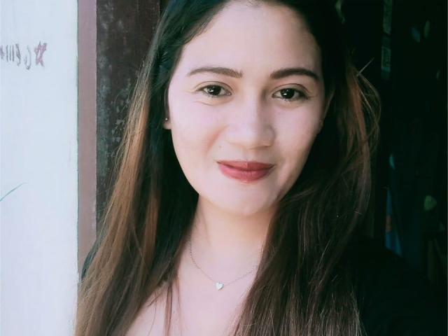 JailyHot has long brown hair, light skin, and is smiling with red lipstick. She's wearing a small heart necklace and a black top, with brown eyes and subtle makeup.