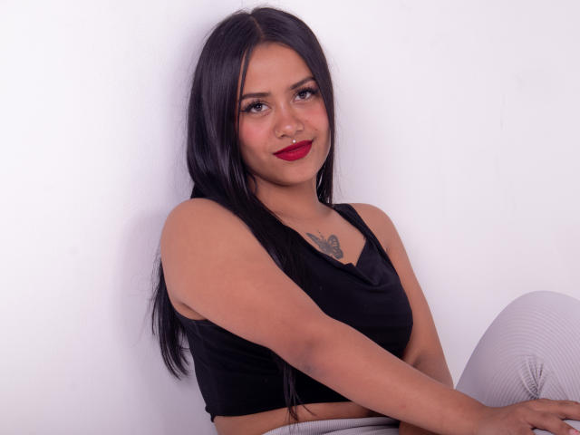 Tarah Brown, con cabello negro largo, lápiz labial rojo, top negro sin mangas, pantalones blancos, pequeña tatuaje de mariposa en el pecho, sentada, sonriendo, con un piercing en la nariz, piel clara, mirando a la cámara..