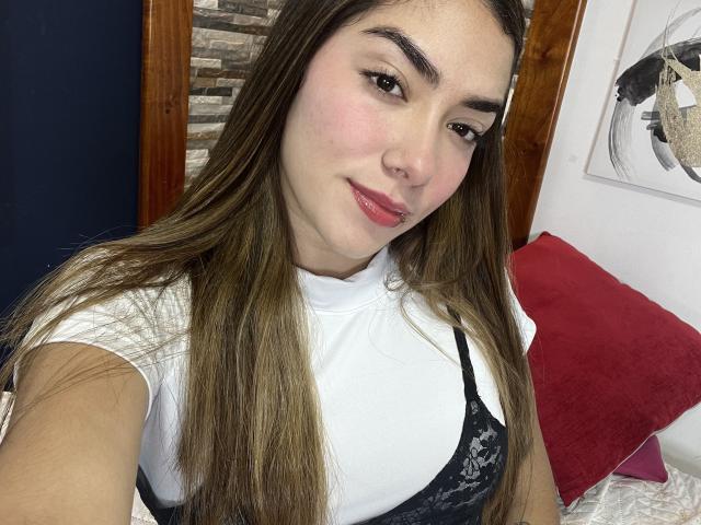 SharonKlart with long brown hair, white shirt, black lace bra, small nose ring, and pink lips, smiling at the camera, showing her tits.