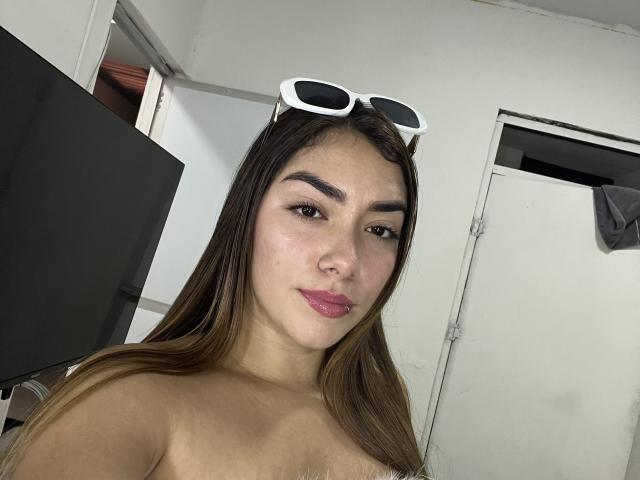 SharonKlart with long brown hair, white sunglasses on head, pink lipstick, and brown eyes, looking at camera, topless, showing small tits, in a white-walled room.