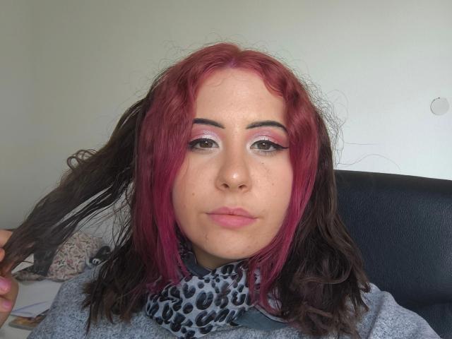 Marizza-hot with red and black hair, pink eyeshadow, and a leopard print scarf, holding her hair, wearing a gray top, and looking at the camera.