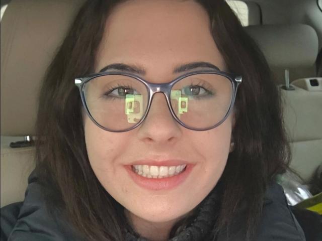 Marizza-hot with dark brown hair, wearing blue glasses, smiling with white teeth. Reflection in glasses shows a green question mark. She's in a car, wearing a dark jacket.