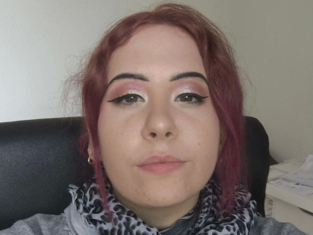 Marizza-hot has red hair, pink eyeshadow, black eyeliner, and a slight smile. She's wearing a gray scarf with black patterns and a small gold earring.