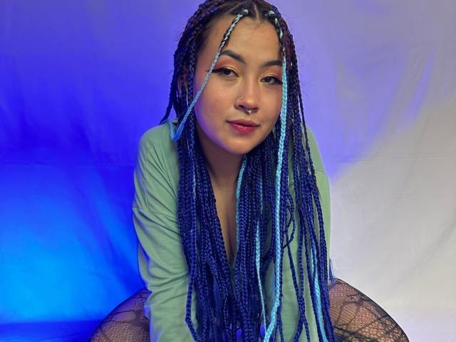 KionI has blue and purple braids, a nose ring, and wears a light green shirt with fishnet stockings, kneeling with a subtle smile, showing a cute face and tits.