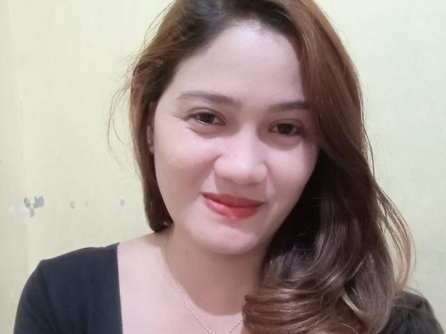 JailyHot has light skin, brown wavy hair, and red lipstick. She's smiling with her eyes slightly closed, wearing a black top and a thin necklace.