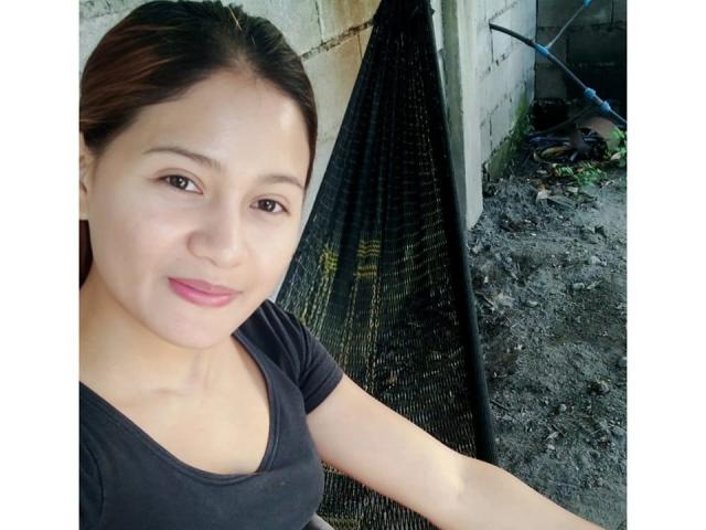JailyHot with light brown skin, black hair, and pink lipstick, wearing a black top, smiling at the camera, standing near a black metal grate on a dirty ground.