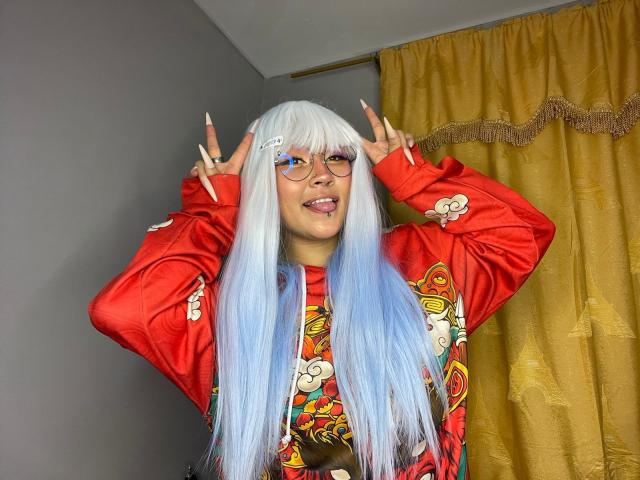 KionI heeft lang wit en blauw haar, een bril en een rood shirt met kleurrijke patronen. Ze toont een vrede-teken met beide handen, met haar tong uit, in een kamer met gele gordijnen..