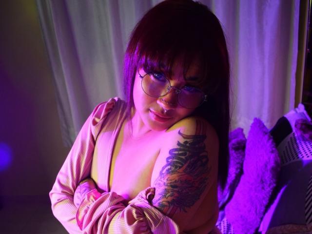 KionI with pinkish-purple hair, glasses, and a nose ring, exposing her tits, has a colorful dragon tattoo on her right shoulder, sitting in a purple-lit room.