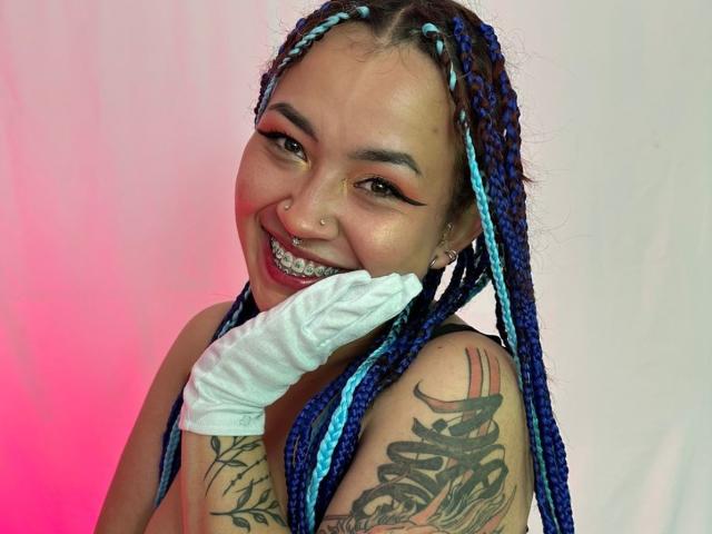 KionI has blue and white braids, a nose ring, and a tattooed arm. She's smiling with white gloves, showing her teeth. Her tits are not visible.