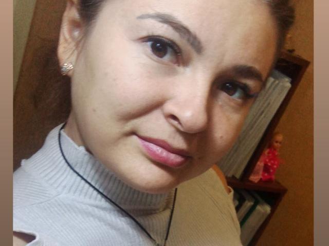 RoxannaSonia's face, showing brown eyes, pink lips, light skin, subtle makeup, and a gray turtleneck, with her brown hair in a ponytail and small diamond earrings.