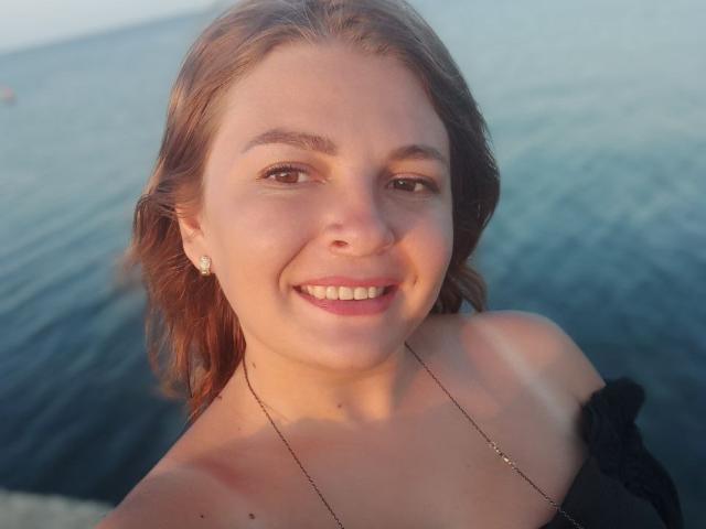 RoxannaSonia has fair skin, brown eyes, and a smile showing white teeth. She's wearing a black off-shoulder top and a gold necklace, with her brown hair down.