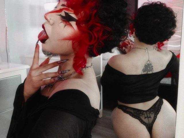 GothVioleth con cabello rizado rojo y negro, top negro con un hombro descubierto, ropa interior de encaje negra, lengua afuera, dedo cerca de la boca, tatuaje en la espalda, mostrando el trasero, reflejo en un espejo. Pechos, trasero, tatuaje, ropa interior..