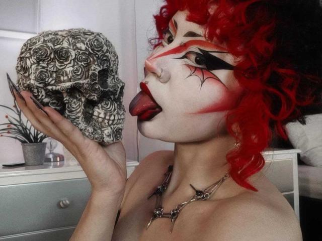 GothVioleth met felrood krullend haar, make-up en zwarte eyeliner, tong uit, likt een schedel met een rozenpatroon, houdt deze vast met één hand, draagt een schedelketting..