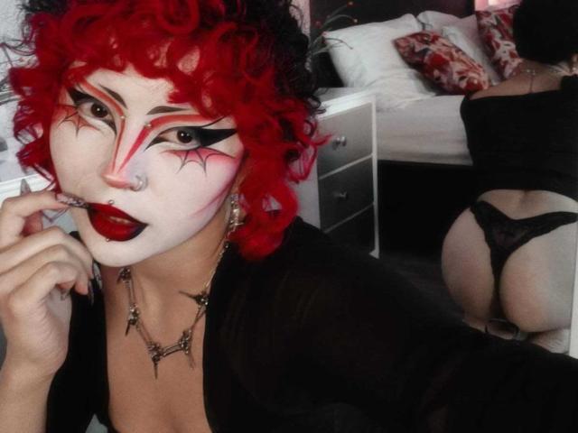 GothVioleth avec des cheveux roux bouclés, du maquillage blanc sur le visage, de l'eye-liner noir, du rouge à lèvres rouge, un anneau au nez, un haut noir, un doigt dans la bouche, montrant ses fesses en culotte en dentelle noire dans un miroir..