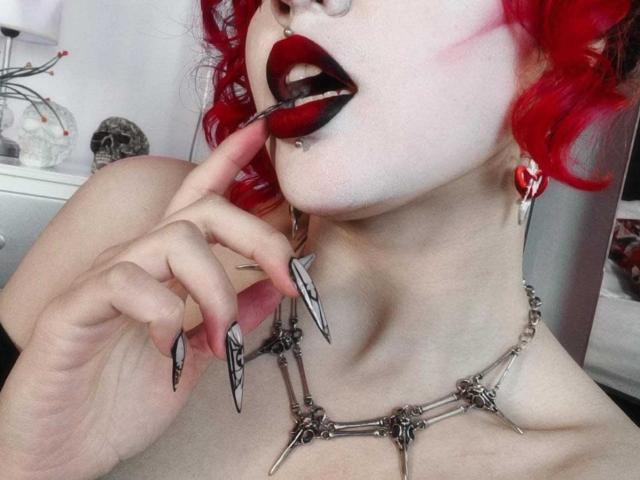 GothVioleth con cabello rizado rojo brillante, piel pálida y lápiz labial negro, chupando una piruleta roja, con un dedo en la boca, usando un collar con púas y uñas largas con puntas negras..