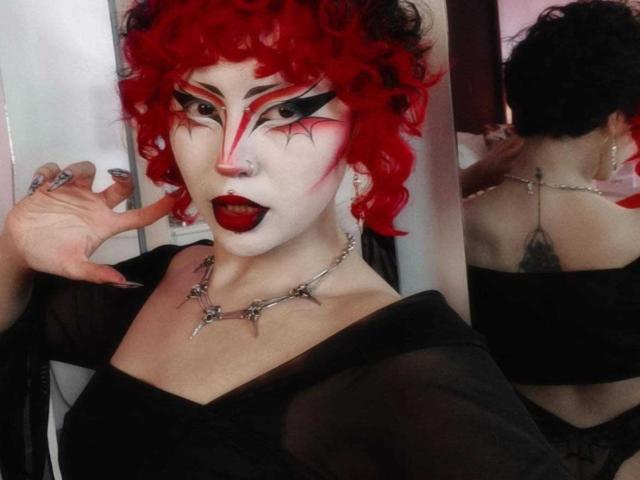 GothVioleth con cabello rizado rojo brillante, maquillaje blanco en la cara, delineador de ojos negro, lápiz labial rojo, vestido negro, collar con púas y tatuaje en la espalda, mostrando uñas cuidadas y un piercing en la nariz..