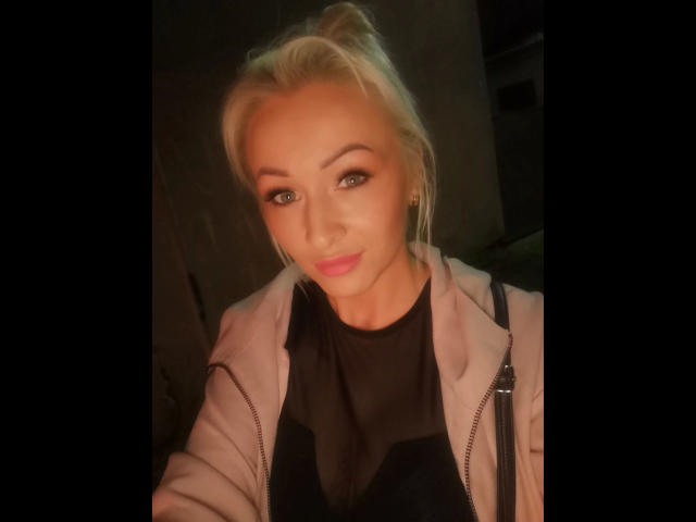 Michaela2015-hot with blonde hair, green eyes, pink lips, wearing a black top and beige jacket, looking at the camera with a slight smile.