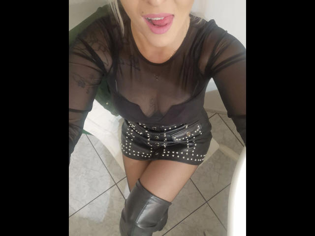 Michaela2015-hot wearing a sheer black top, showing her small tits, and a black leather skirt with studs, sitting with her knee up, wearing black knee-high boots.