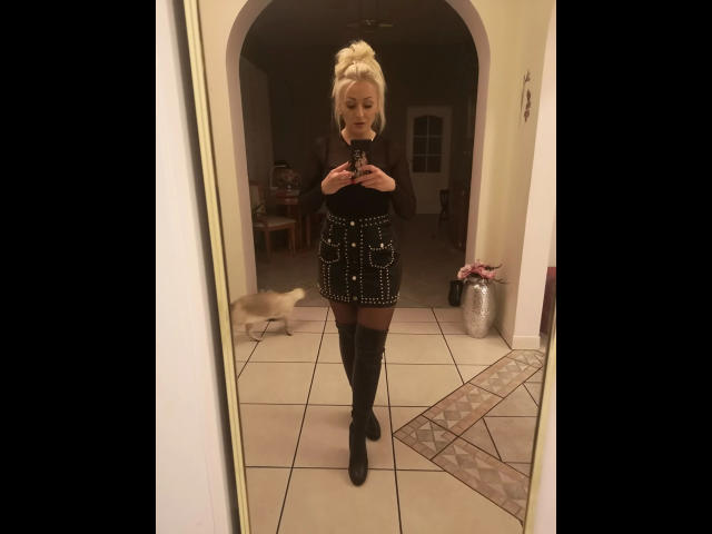Michaela2015-hot, blonde, black turtleneck, black skirt with studs, black thigh-high boots, standing in front of mirror, holding phone, cat blurred in background, tiled floor.