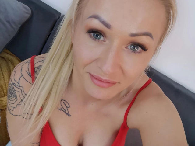 Michaela2015-hot with blonde hair, blue eyes, red lipstick, red bra, rose tattoo on right arm, small tattoo below collarbone, smiling, looking at camera.