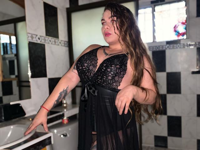 BlueSofy with long brown hair, wearing black lace lingerie, has large tits and a tattoo on her right arm, standing in a bathroom with black and white tiles.