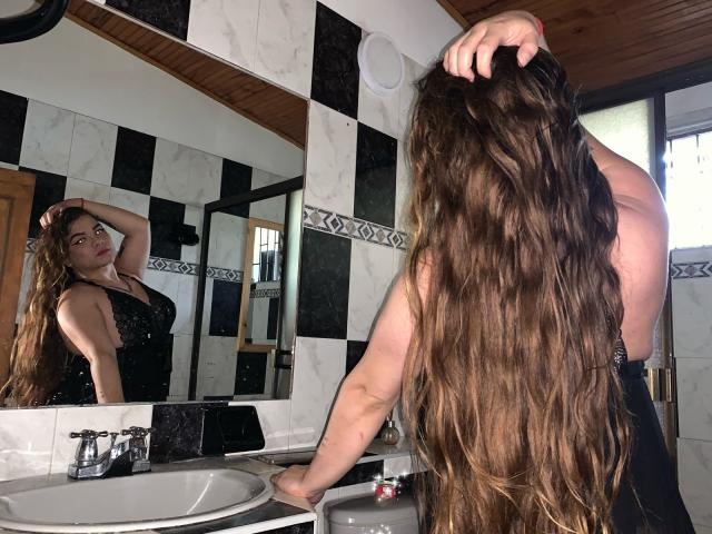 BlueSofy with long brown wavy hair, back to camera, hand in hair, black dress, reflection in mirror, white and black tile bathroom, wooden ceiling, hand on sink.