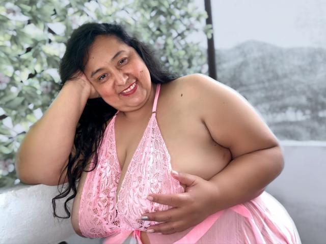 TistMature has large, round tits in a pink lace halter top, smiling with black hair, one hand on her head, and the other touching her chest.