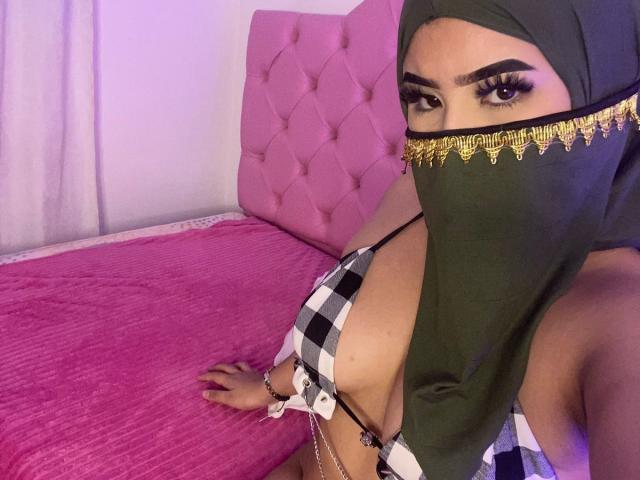 MiaHadid with green headscarf, checkered top, gold chain, and mask, showing large tits, pink bed, and pink headboard, looking at camera with dramatic eye makeup.