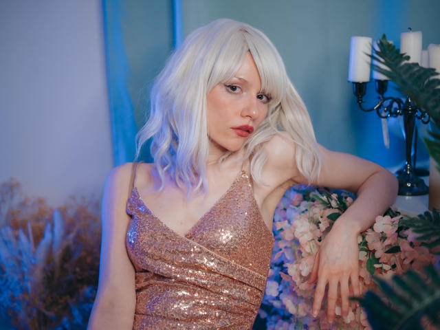 BrendaLovex with white hair, wearing a sparkling pink dress, has small tits, a cute face, and a sexy, relaxed pose, leaning on flowers with a candle in the background.