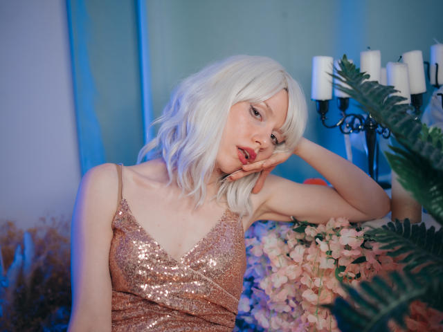 Brenda Lovex, mit weißem Haar, trägt ein glitzerndes pinkes Kleid und legt ihren Kopf in ihre Hand, wobei sie ihre Brüste zur Schau stellt. Sie befindet sich in einem blau beleuchteten Raum, umgeben von Blumen und Kerzen..
