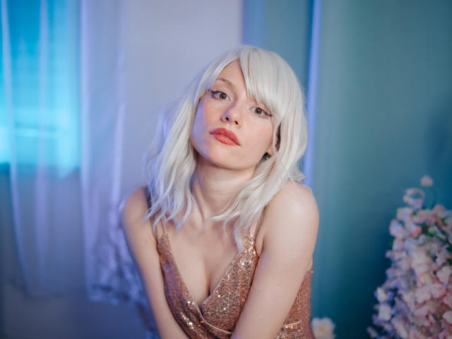 Brenda Lovex mit blonden Haaren, in einem glitzernden pinken Kleid, hat kleine Brüste, lehnt sich vor, hat rote Lippen und einen leicht geneigten Kopf, in einem blau beleuchteten Raum mit Blumen..