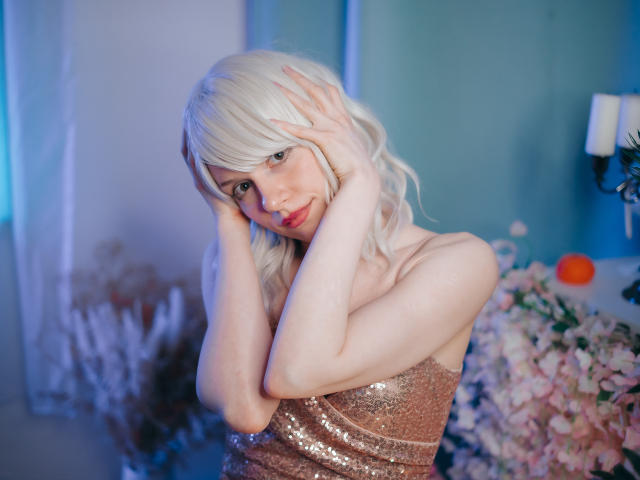BrendaLovex mit einer platinblonden Perücke, Hände im Haar, trägt ein glitzerndes pinkes Kleid, wirkt verführerisch mit roten Lippen, heller Haut und kleinen Brüsten, in weichem blauen Licht..