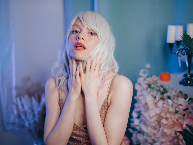 BrendaLovex with pale skin, white wavy hair, pink lipstick, and a sparkly brown dress, hands touching her neck, looking up with a slight smile. Soft blue and pink lighting.