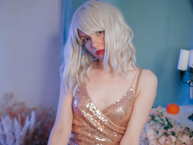BrendaLovex with blonde wig, pink sequined dress, and pale skin, has small tits, pouty pink lips, and a shy, sultry look, standing in a softly lit, floral setting.