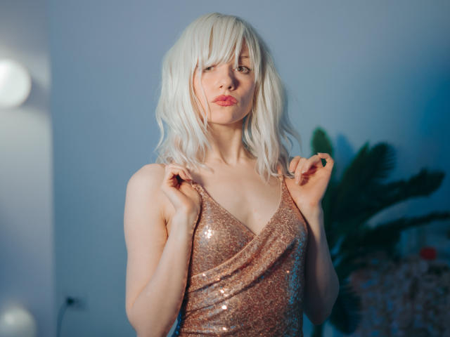 Brenda Lovex mit platinblondem Haar, trägt ein schimmerndes, roségoldenes, paillettenbesetztes Kleid, steht da, die Hände in der Nähe ihrer Schultern, helle Haut, sinnliche, rosafarbene Lippen, blaugrauer Hintergrund, weiches Licht..